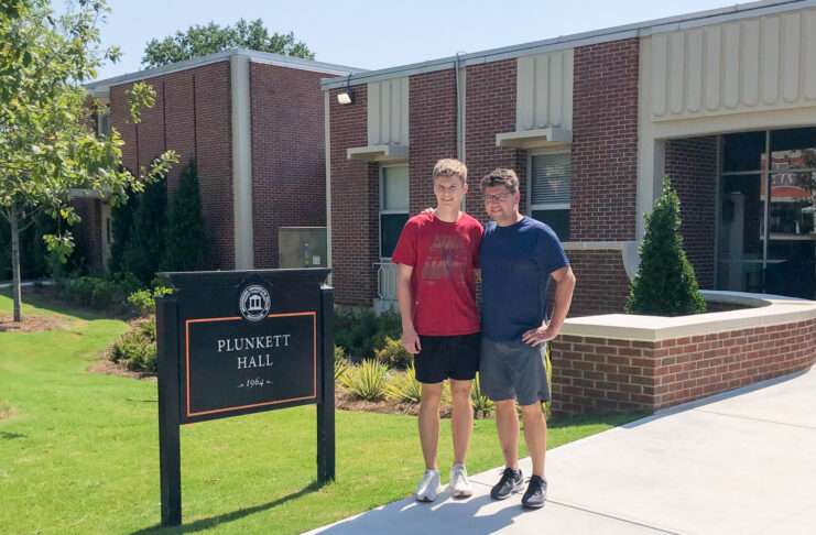 Father and son stand outside in front of a 1960s style, brick building and next to a black sign that says Plunkett Hall.