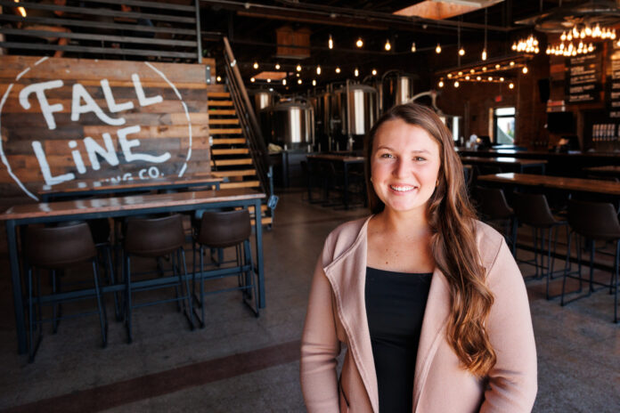 Woman wearing a black shirt and tan sweater smiling in the foreground with "Fall Line Brewing Co." sign in the background inside a brewpub.