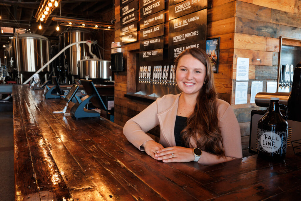 Woman wearing a black shirt and tan sweater smiling at a bar in Fall Line Brewery, with brewing tanks visible in the background and a menu board listing beer options.