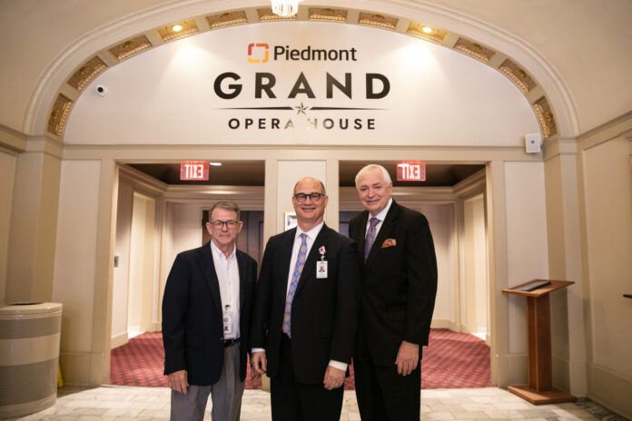 Three men stand in a grand lobby with the words "Piedmont Grand Opera House" printed on the wall behind them.