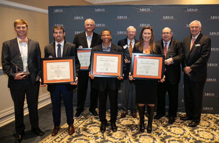 A group of eight individual stands in front of a Mercer backdrop, holding framed certificates and awards.