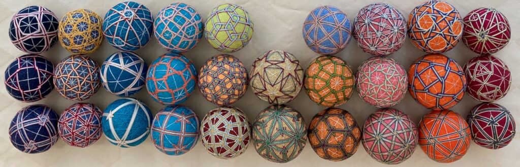 A set of colorful, intricately embroidered balls, likely handcrafted in a traditional Japanese Temari style. Each ball is adorned with geometric patterns and vibrant threadwork, showcasing a variety of designs such as stars, polygons, and intersecting lines in different color combinations.