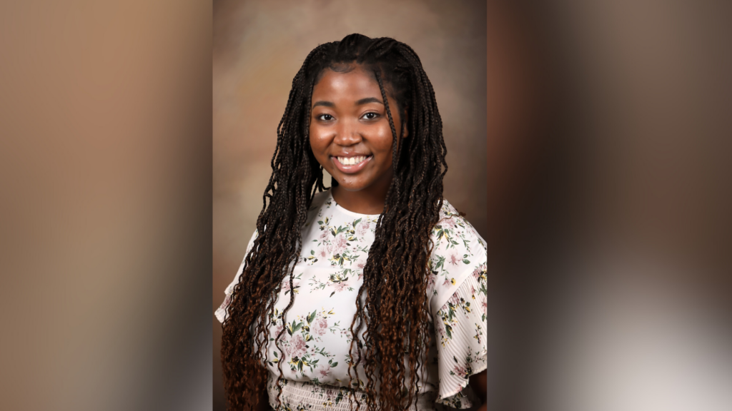 A woman smiling and wearing a floral-patterned top with long braided hair. The background is a soft, neutral, and slightly blurred color gradient