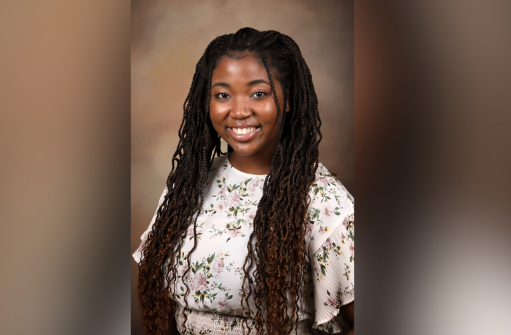 A woman smiling and wearing a floral-patterned top with long braided hair. The background is a soft, neutral, and slightly blurred color gradient