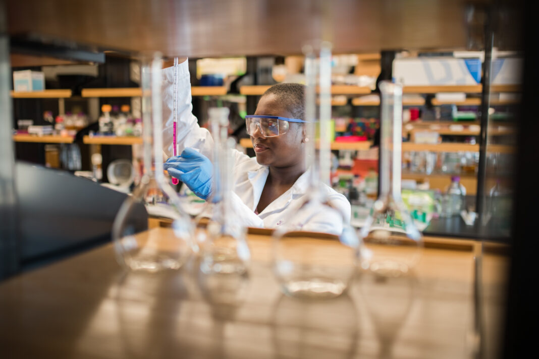 Person in a laboratory setting, wearing safety goggles, gloves, and a lab coat. They are working with a pipette or similar tool. Various glass laboratory equipment, such as beakers or flasks, can be seen in the foreground, slightly out of focus. The background is a typical research lab, with shelves containing scientific instruments and materials.