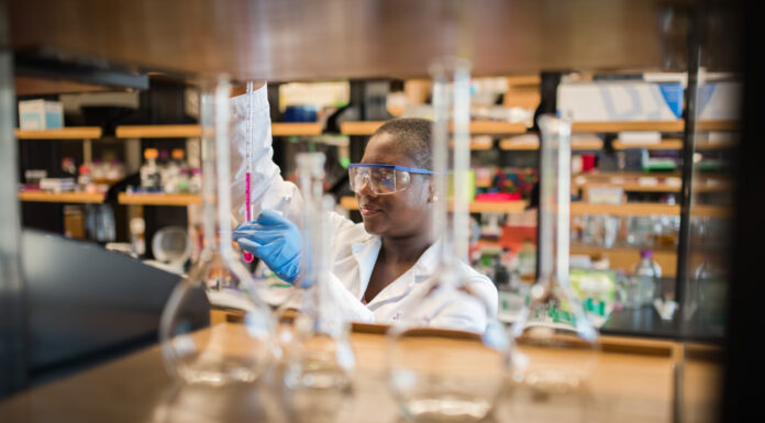 Mercer receives $116,000 NSF grant for material science research Person in a laboratory setting, wearing safety goggles, gloves, and a lab coat. They are working with a pipette or similar tool. Various glass laboratory equipment, such as beakers or flasks, can be seen in the foreground, slightly out of focus. The background is a typical research lab, with shelves containing scientific instruments and materials.