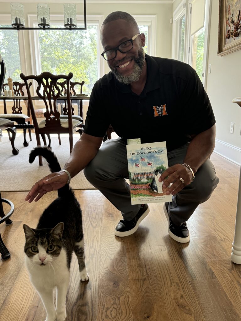 A man crouches to pet a striped cat while holding a children's book titled "Veto, the Governor's Cat."