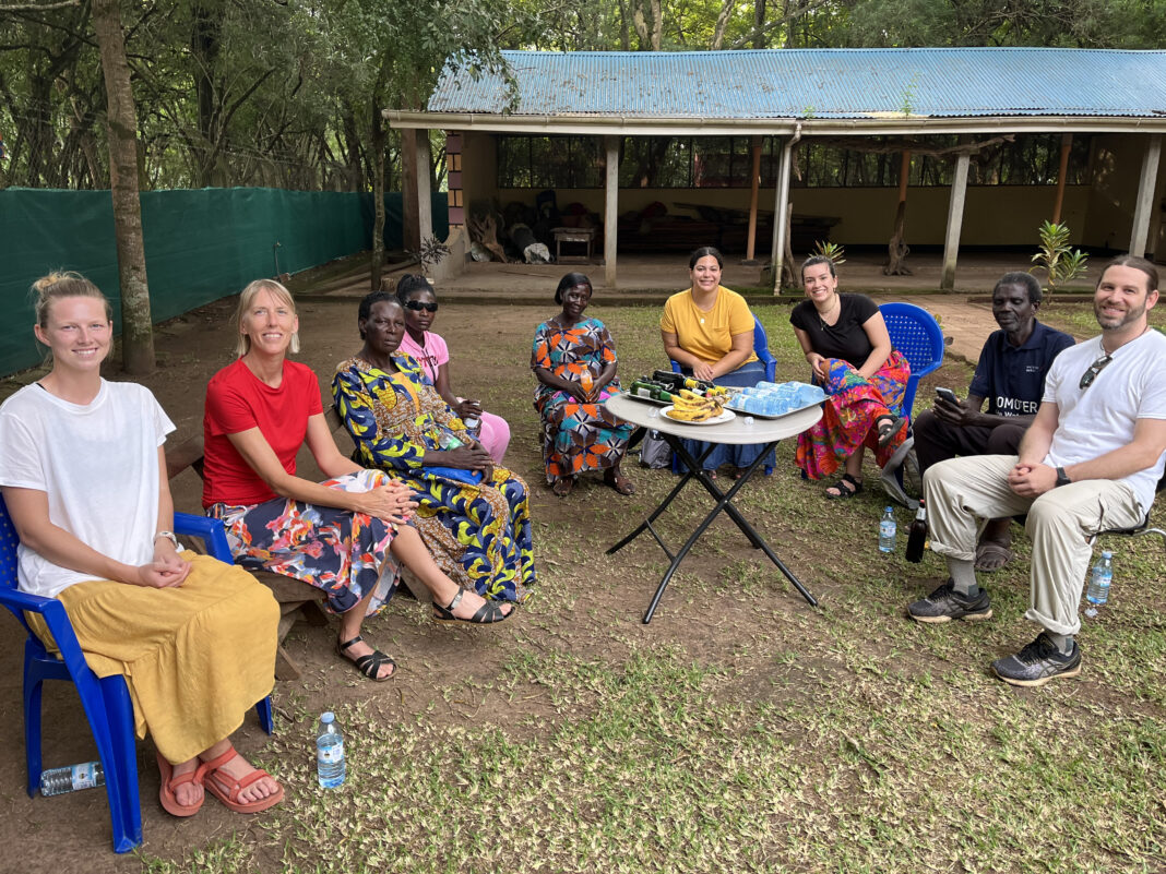 Group of eight people, consisting of both men and women, are seated outdoors in a semicircle on plastic chairs. The group includes individuals of different cultural backgrounds. Some people are wearing brightly patterned traditional attire, while others are dressed casually in modern clothing. In the center of the group, there is a small table with snacks, including bananas and bottled water. Behind them, a simple structure with a corrugated metal roof and open walls is visible, surrounded by trees and greenery.