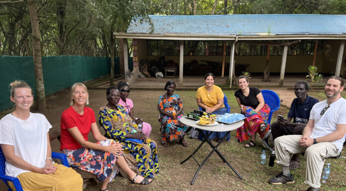 Anthropology major marks inaugural year Group of eight people, consisting of both men and women, are seated outdoors in a semicircle on plastic chairs. The group includes individuals of different cultural backgrounds. Some people are wearing brightly patterned traditional attire, while others are dressed casually in modern clothing. In the center of the group, there is a small table with snacks, including bananas and bottled water. Behind them, a simple structure with a corrugated metal roof and open walls is visible, surrounded by trees and greenery.