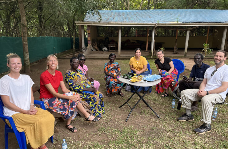 Group of eight people, consisting of both men and women, are seated outdoors in a semicircle on plastic chairs. The group includes individuals of different cultural backgrounds. Some people are wearing brightly patterned traditional attire, while others are dressed casually in modern clothing. In the center of the group, there is a small table with snacks, including bananas and bottled water. Behind them, a simple structure with a corrugated metal roof and open walls is visible, surrounded by trees and greenery.