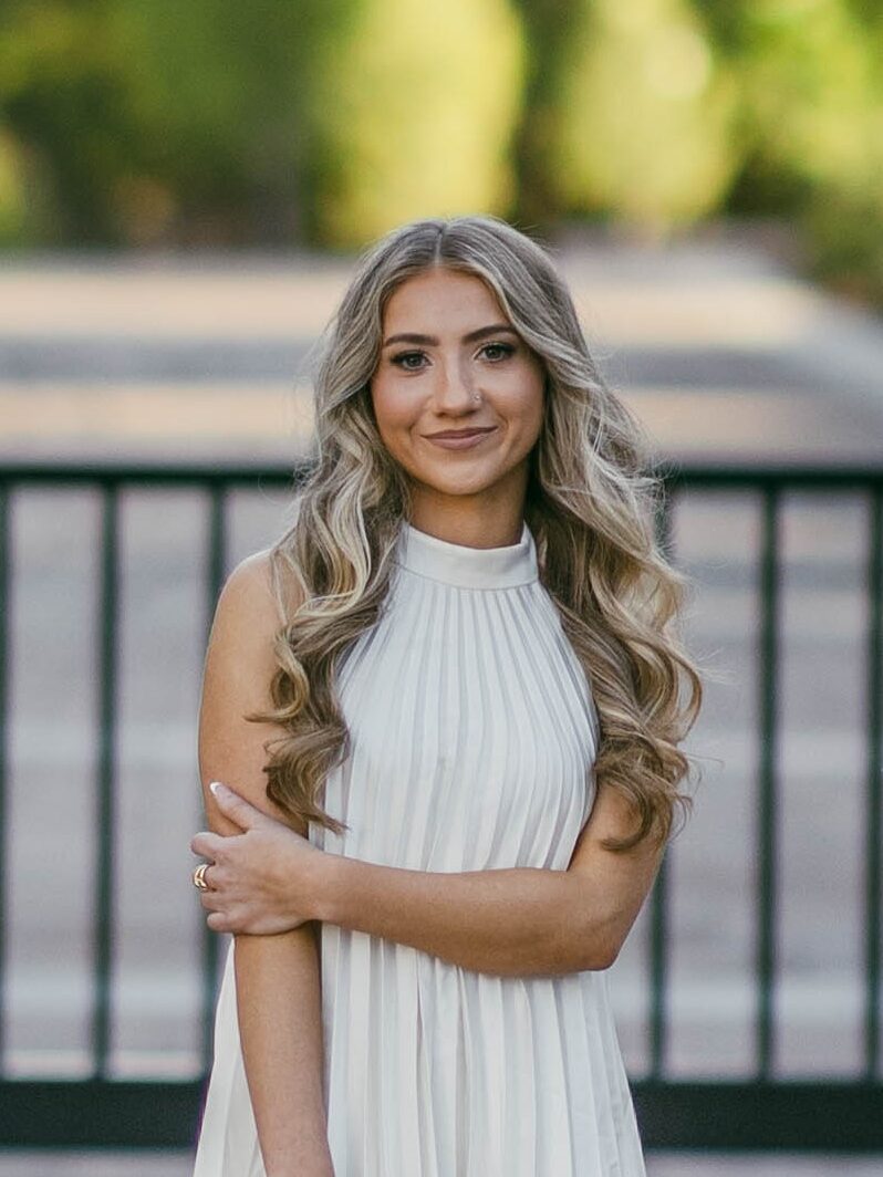 Professional headshot of a young woman standing outdoors. She has long, light blonde hair and is smiling warmly at the camera. She is wearing a white dress The background shows blurred greenery and a fence with soft lighting. 