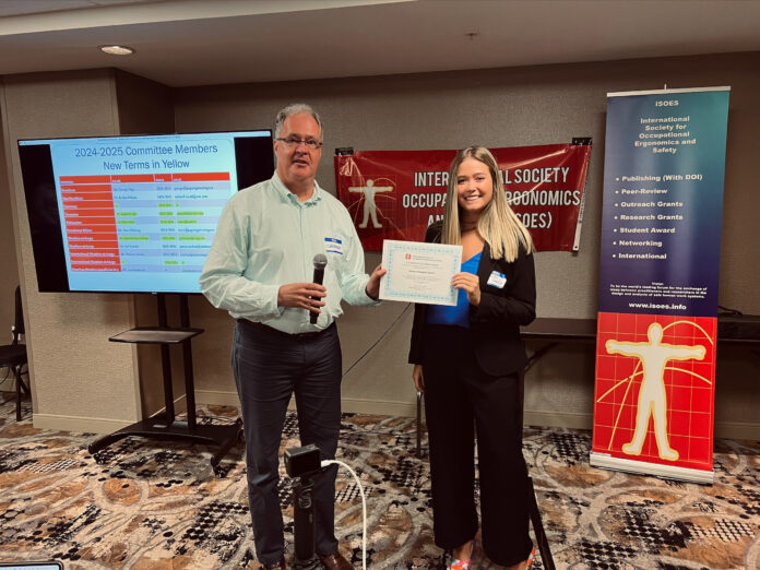 Two individuals at a conference, standing side by side. The man on the left is holding a microphone and smiling, while the woman on the right is holding a certificate. Behind them is a banner for the "International Society for Occupational Ergonomics and Safety" and a presentation screen showing the "2024-2025 Committee Members" list, with new members highlighted in yellow. The banner lists ISOES initiatives, including "Publishing (With DOI)," "Peer-Review," "Outreach Grants," and more. The setting appears to be indoors, in a conference room.