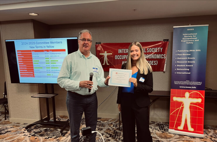 Two individuals at a conference, standing side by side. The man on the left is holding a microphone and smiling, while the woman on the right is holding a certificate. Behind them is a banner for the 