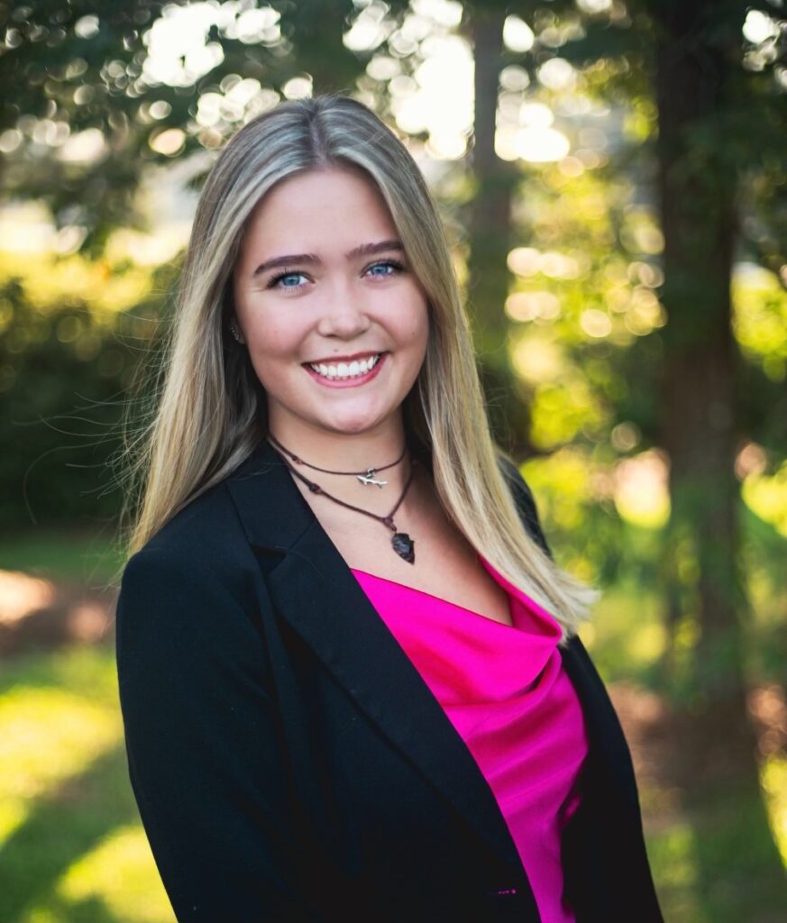 Professional headshot of a young woman standing outdoors. She has long, light blonde hair and is smiling warmly at the camera. She is wearing a black blazer over a bright pink top and a necklace with a pendant. The background shows blurred greenery with soft lighting.