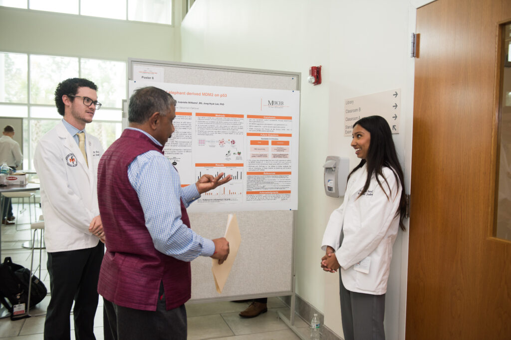 Three individuals are engaged in a discussion next to a scientific poster presentation in a brightly lit room.