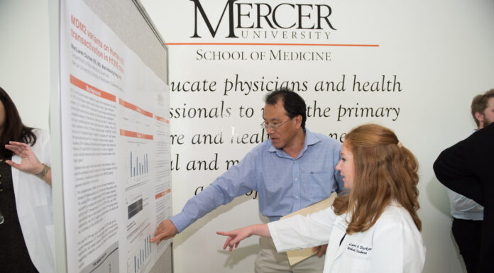 Together for a Cure: Mercer medical students contribute to cancer research A Mercer medical student in a lab coat points at a poster next to a man also pointing at the poster.