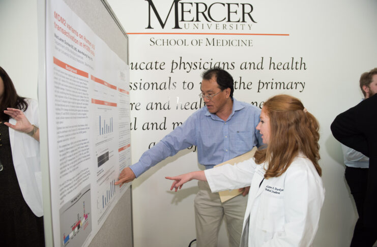 A Mercer medical student in a lab coat points at a poster next to a man also pointing at the poster.