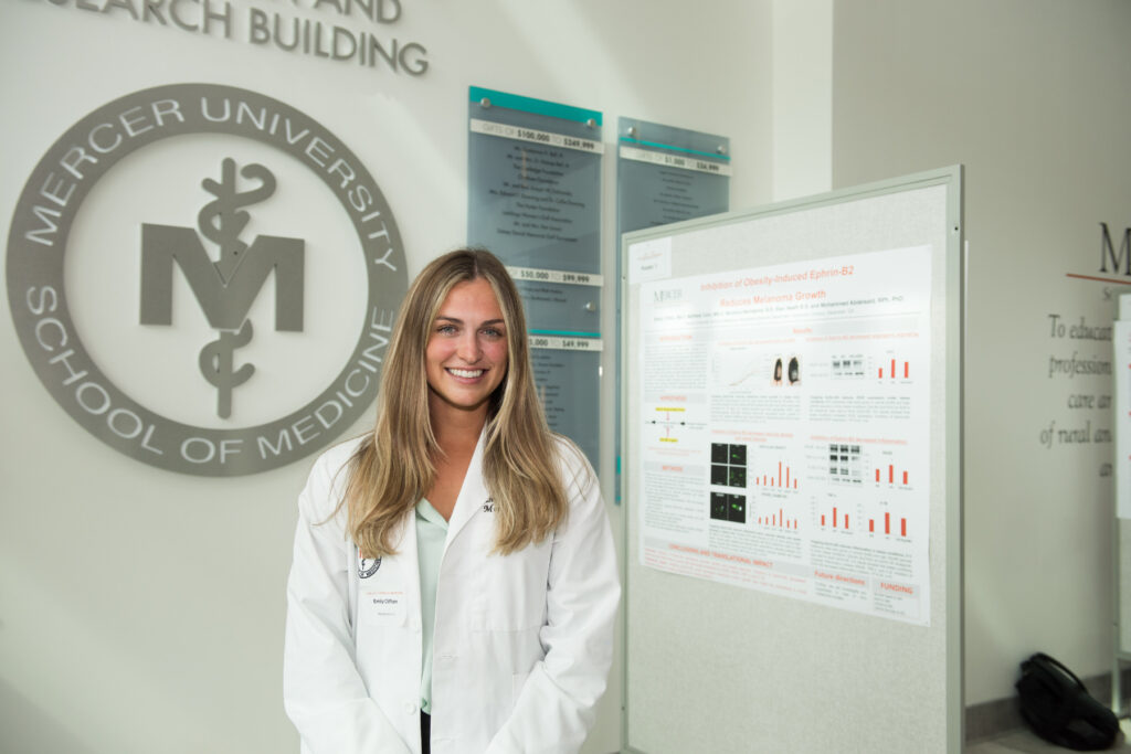 A person in a white lab coat stands smiling in front of a research poster at Mercer University School of Medicine.