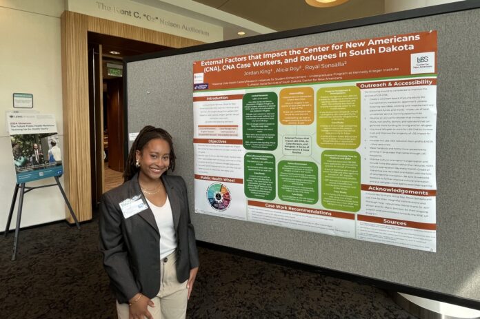 A woman is standing next to a presentation board. The title of the board reads: "External Factors that Impact the Center for New Americans (CNA), CNA Case Workers, and Refugees in South Dakota." The board outlines several sections such as "Introduction," "Objectives," "Outreach & Accessibility," and "Case Work Recommendations." The woman is smiling, wearing a grey blazer, white top, and khaki pants.