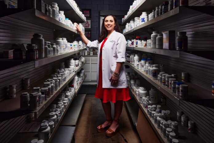 A woman in a red dress and a white lab coat stands in between two shelves of prescription medicines and smiles. She is illuminated by light, with dark shadows around her.