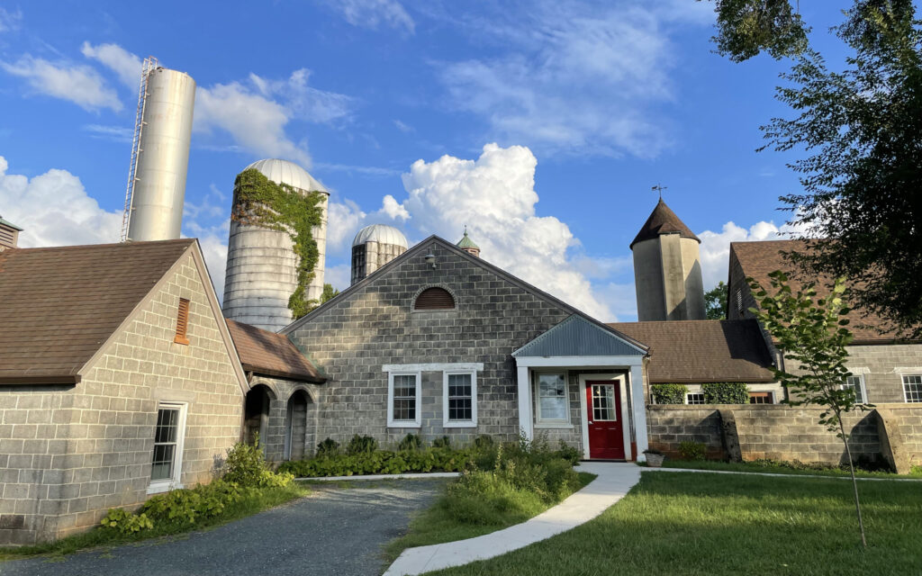 A rustic farm-like setting featuring multiple buildings with gray stone or cinder block exteriors. The structures have pitched brown roofs and are surrounded by silos, including one with ivy growing on it. A bright red door stands out on one of the buildings, and a curved gravel pathway leads up to the entrance. A clear blue sky with scattered clouds serves as the backdrop, while greenery and trees frame the scene.