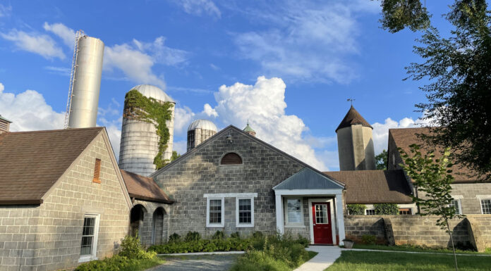Writing and design faculty delve into creative projects during summer residency A rustic farm-like setting featuring multiple buildings with gray stone or cinder block exteriors. The structures have pitched brown roofs and are surrounded by silos, including one with ivy growing on it. A bright red door stands out on one of the buildings, and a curved gravel pathway leads up to the entrance. A clear blue sky with scattered clouds serves as the backdrop, while greenery and trees frame the scene.
