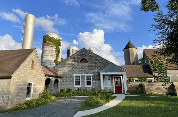 A rustic farm-like setting featuring multiple buildings with gray stone or cinder block exteriors. The structures have pitched brown roofs and are surrounded by silos, including one with ivy growing on it. A bright red door stands out on one of the buildings, and a curved gravel pathway leads up to the entrance. A clear blue sky with scattered clouds serves as the backdrop, while greenery and trees frame the scene.