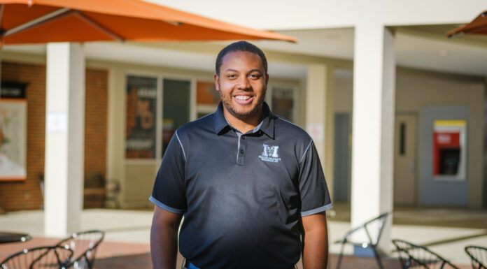 New housing director focused on providing ‘world-class experience’ for students A man wearing a black Mercer polo shirt stands in a courtyard with tables, chairs and an orange umbrella visible. A building is in the background.