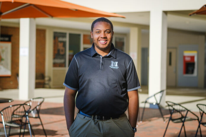 A man wearing a black Mercer polo shirt stands in a courtyard with tables, chairs and an orange umbrella visible. A building is in the background.