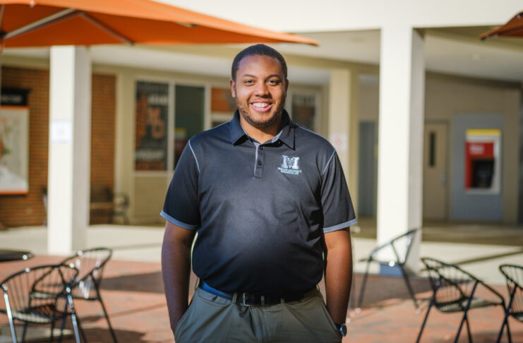 A man wearing a black Mercer polo shirt stands in a courtyard with tables, chairs and an orange umbrella visible. A building is in the background.