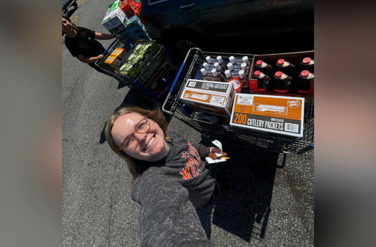 Person smiling at the camera, pushing a shopping cart full of groceries including bottled beverages and cutlery packets in a sunny parking lot.