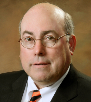 Portrait of Richard A. “Doc” Schneider wearing glasses, a dark suit, and an orange and black tie.