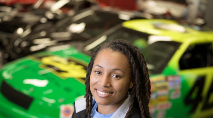 Mercer alumna is a user experience designer for General Motors A woman wearing black jacket and light blue shit is pictured, with brightly colored race cars in the background behind her.
