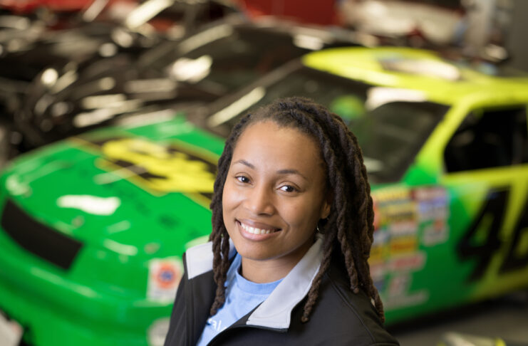 A woman wearing black jacket and light blue shit is pictured, with brightly colored race cars in the background behind her.