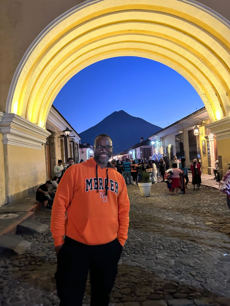 A man in an orange Mercer sweatshirt and black pants stands under an illuminated archway on a cobblestone street in the evening, with a mountain in the background.