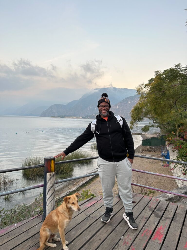 A man in a knit hat, black jacket and khakis stands on a wooden platform with a dog beside him and mountains and a body of water behind him.