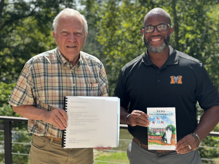 A man in a checkered button-down shirt holds a spiralbound book while a man in a Mercer University polo holds a book titled "Veto, the Governor's Cat."