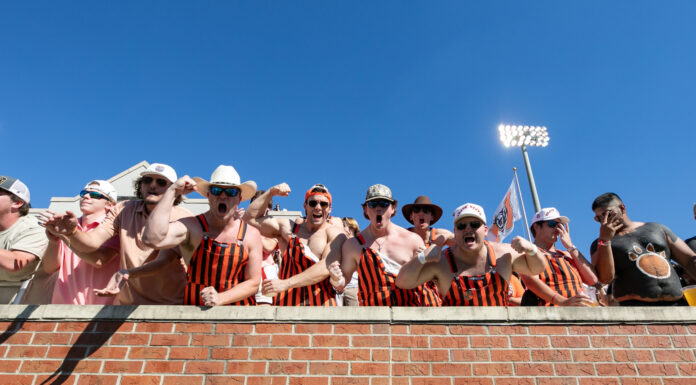 3 things to know about Mercer Homecoming football game day: Nov. 2, 2024 Enthusiastic football fans in matching striped orange-and-black outfits and white hats cheer from stadium stands on a sunny day.