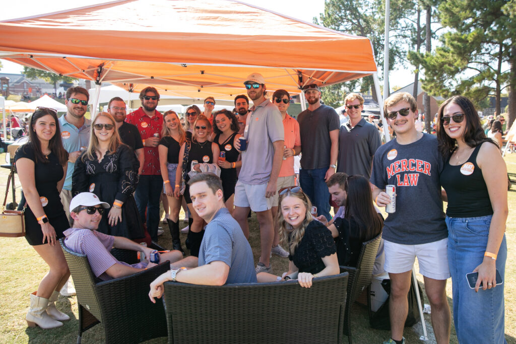 A group of people enjoying an outdoor tailgate under a tent. Some individuals are seated while others stand, all appear to be socializing and smiling on a sunny day.