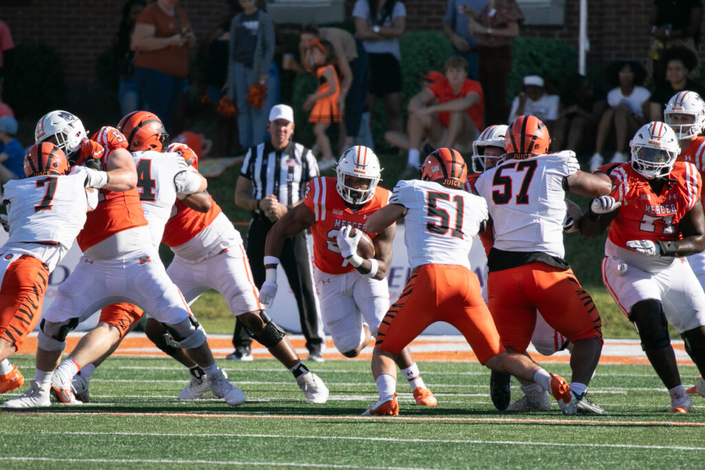 Players from the Mercer Bears and Princeton Tigers in action during a college football game, with a Mercer player charging with the ball while others from both teams engage around them on the field.