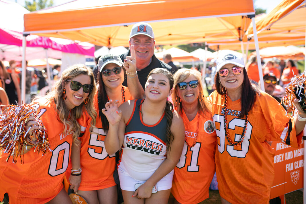 Group of six enthusiastic Mercer University fans wearing orange team apparel and accessories, posing together at a daytime outdoor event.