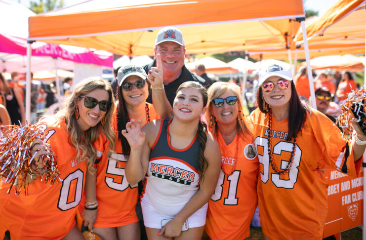 Group of six enthusiastic Mercer University fans wearing orange team apparel and accessories, posing together at a daytime outdoor event.