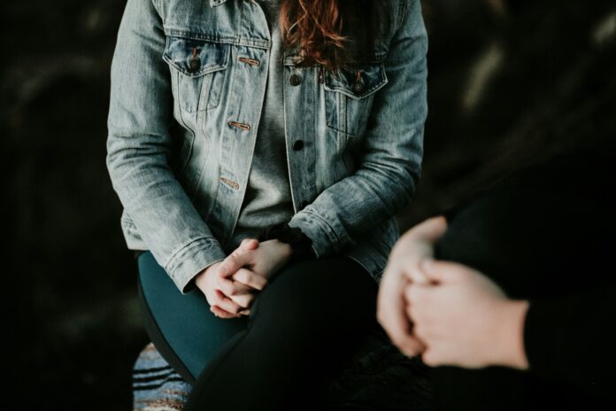 Woman wearing jean jacket with hands clasped on her lap.