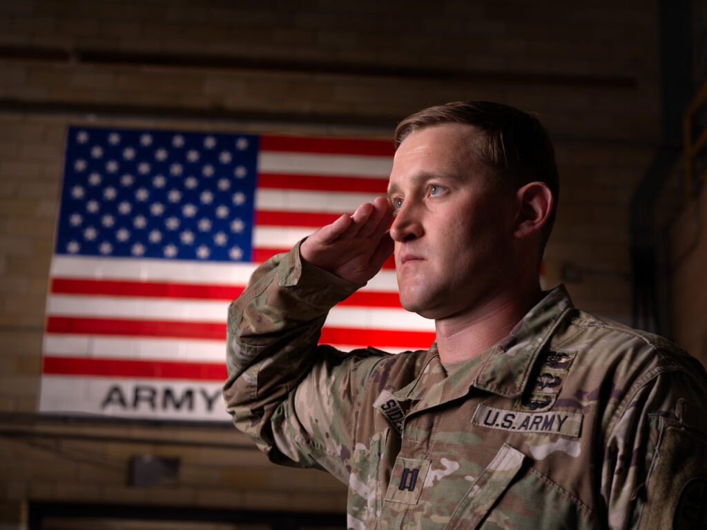 A man in an Army uniform looks ahead while saluting, with the American flag on the wall behind him.