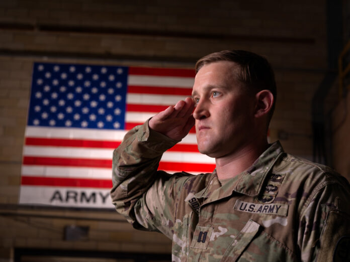 A man in an Army uniform looks ahead while saluting, with the American flag on the wall behind him.