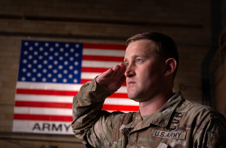 A man in an Army uniform looks ahead while saluting, with the American flag on the wall behind him.