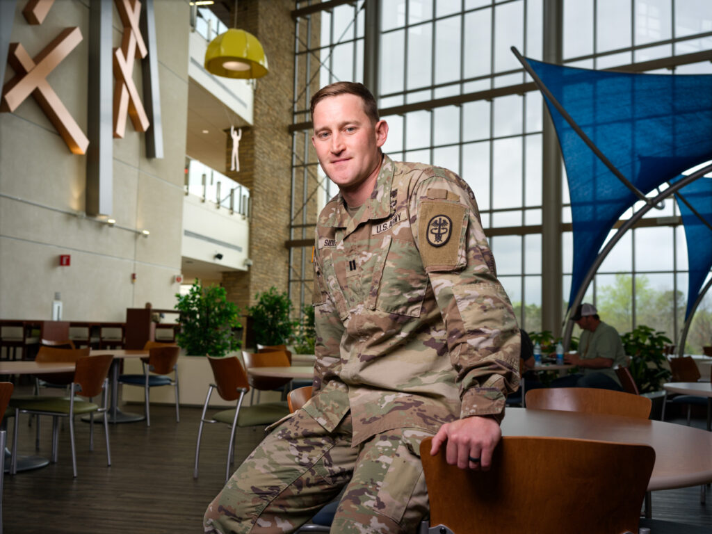 A man in an Army uniform sits on a tables and smiles, with other tables and chairs behind him in an open room with windows covering the back wall.