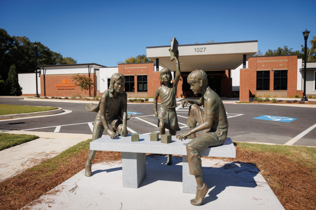 Bronze sculpture of three figures, two on a bench and one holding up a book, in front of Roberts Academy.