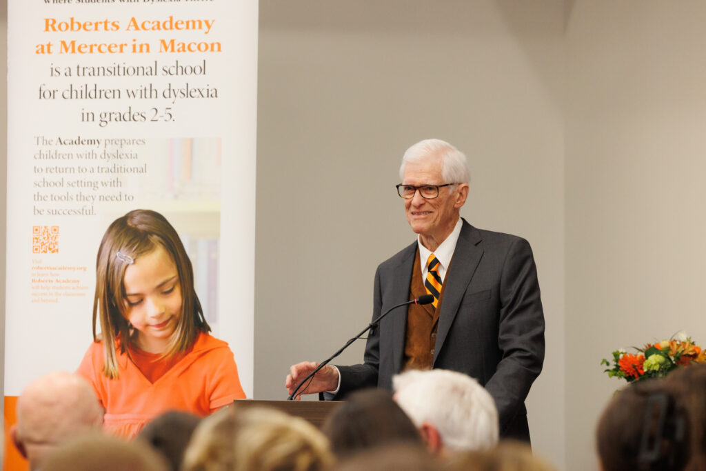 An individual is speaking at Roberts Academy at Mercer in Macon, standing at a podium with attentive audience members in the foreground. A promotional poster beside the speaker highlights the academy's focus on children with dyslexia in grades 2-5.