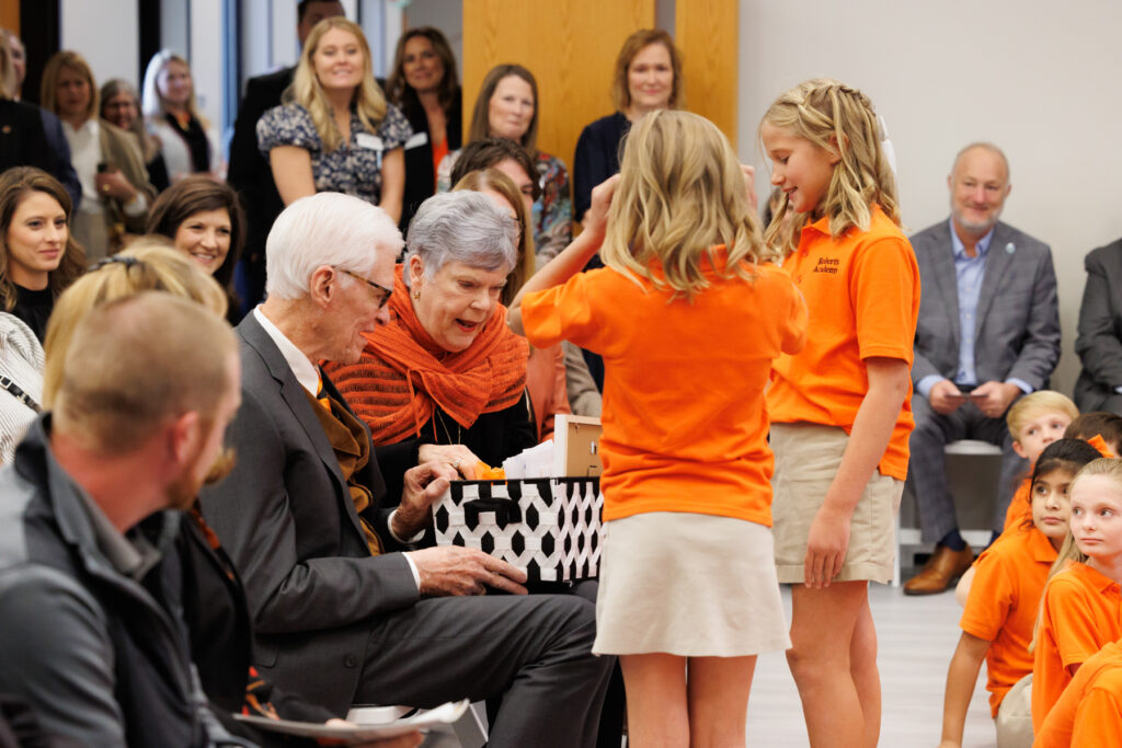 Two children in orange shirts present a gift to an older couple seated in a room filled with onlookers.